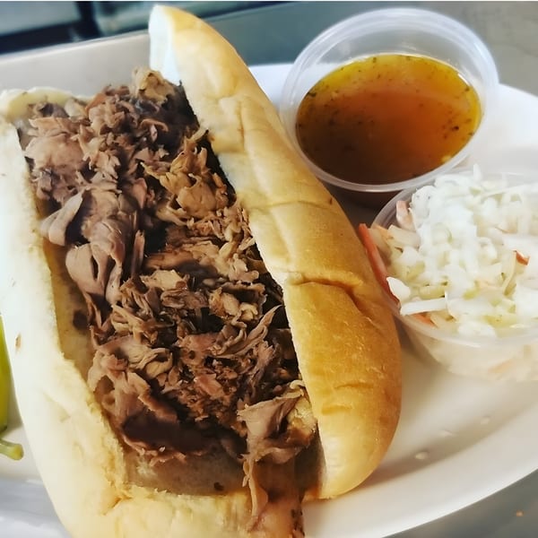 A beef sandwich with a side of coleslaw and a cup of au jus on a white plate.